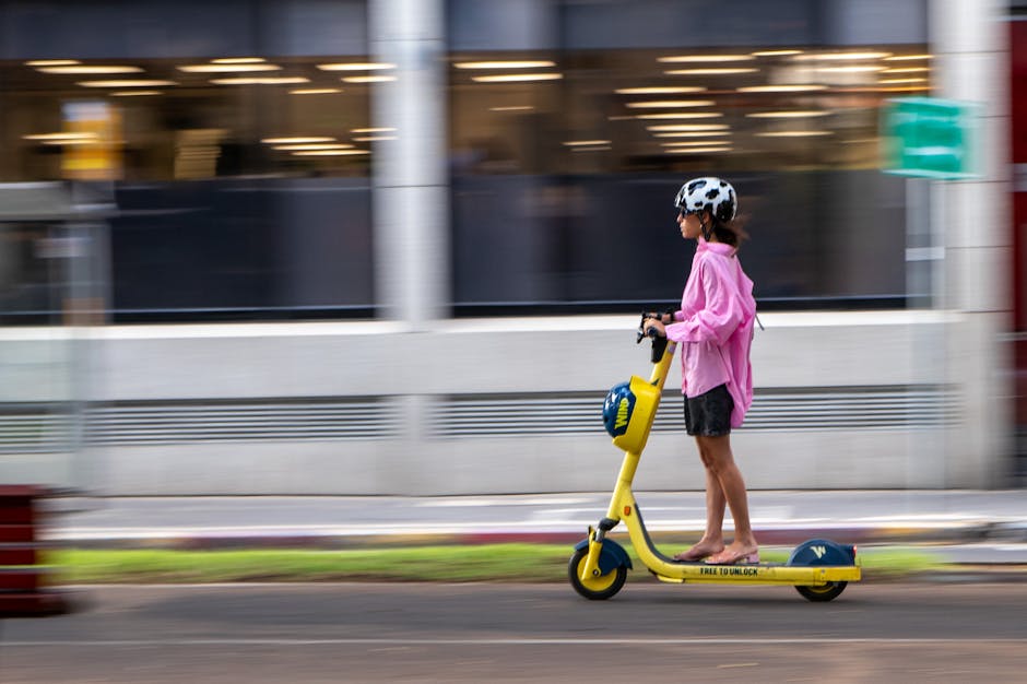 Woman in pink jacket rides a yellow electric scooter on a city street. Blurred motion effect.