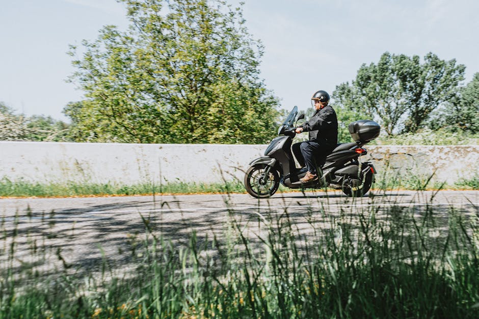 A man enjoys a scenic ride on a motor scooter along a countryside road surrounded by lush green trees.